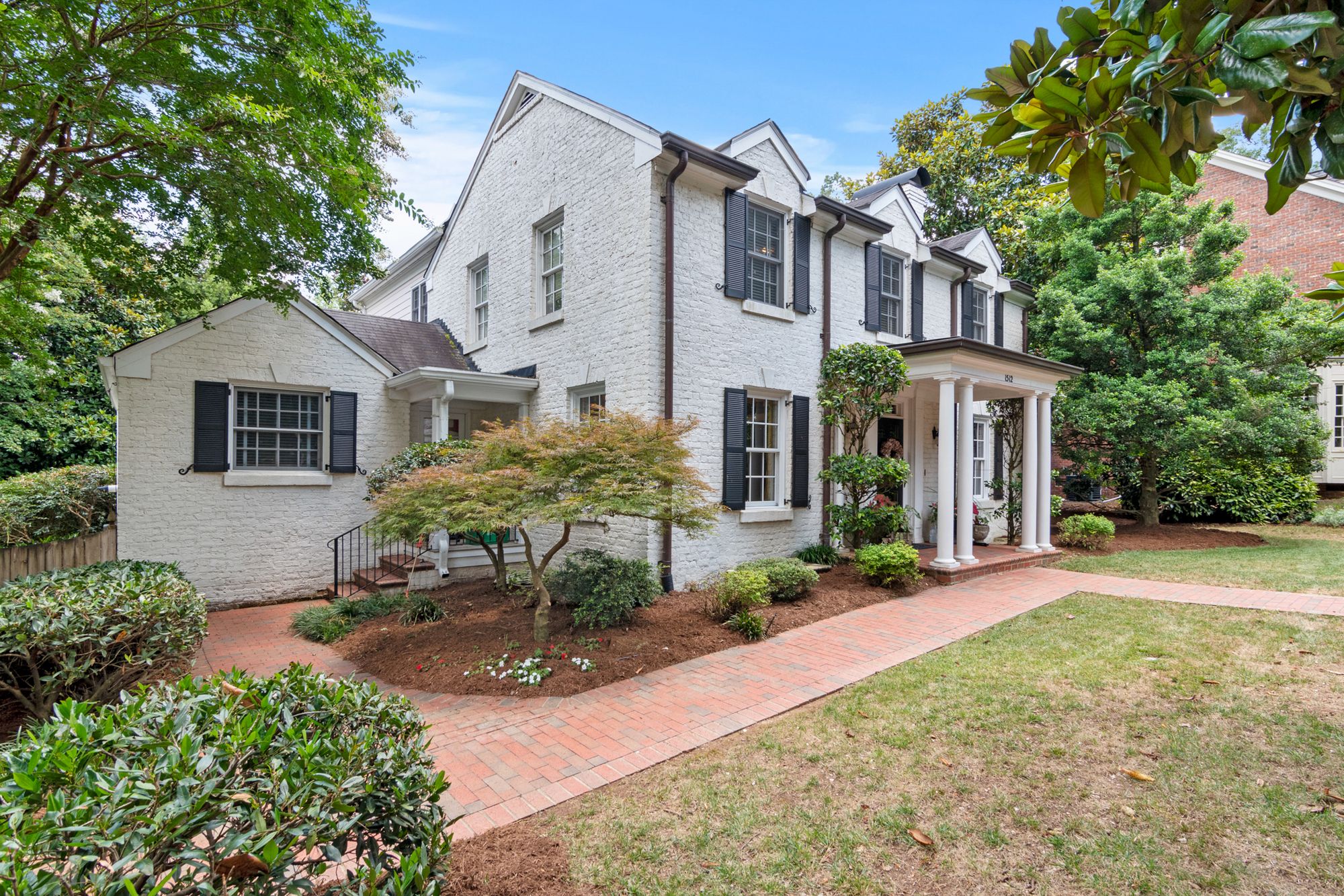 Two-story white brick residence with dark shutters and mature landscaping